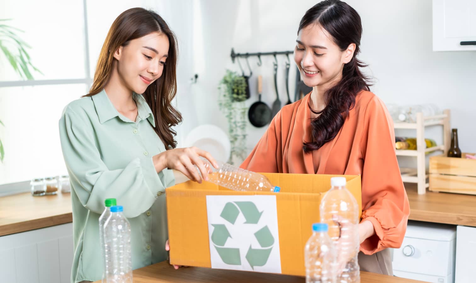 Two women sort plastic bottles into a box in a bright kitchen, promoting recycling efforts