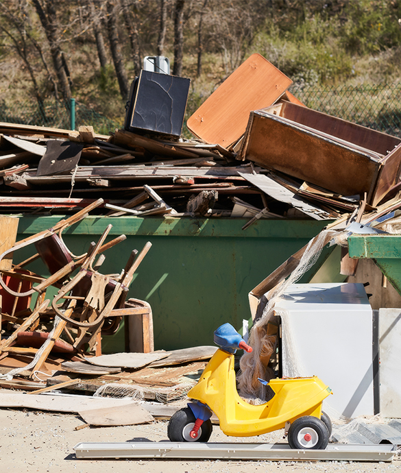 Yellow childs tricycle in front of large pile of discarded furniture and debris by dumpster.